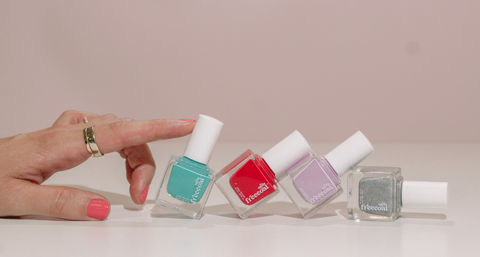Hand with coral manicure and gold rings nudging four tilted nail polish bottles—teal, red, lavender, and silver glitter—on a pale pink surface