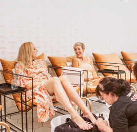Two women laughing in a bright pedicure lounge while a technician gives a foot treatment in a modern nail spa