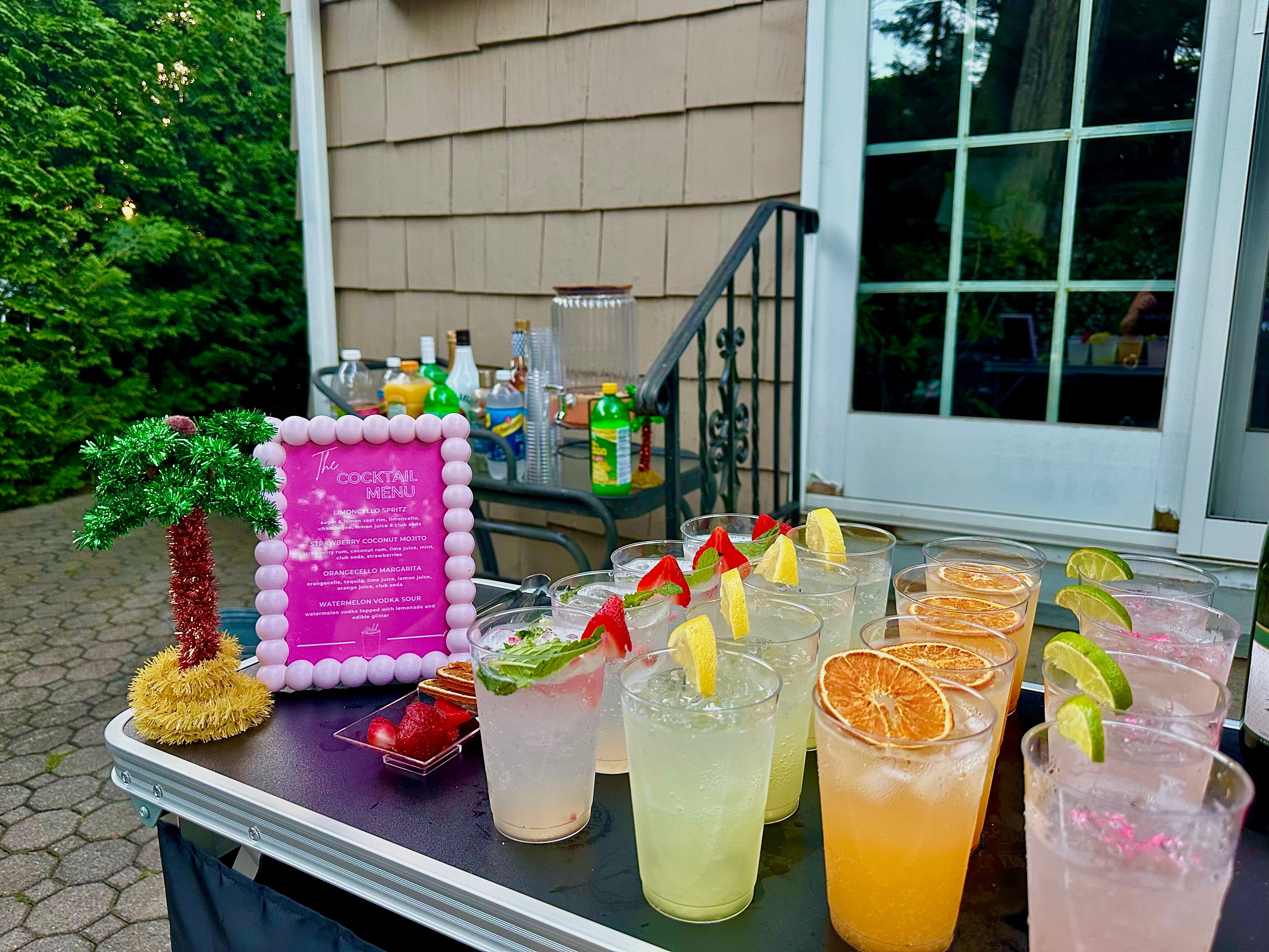 Outdoor backyard patio cocktail table with rows of colorful garnished drinks in plastic cups—lemon, lime, dried orange slices and strawberries—next to a pink-framed cocktail menu and a small tinsel palm tree, ready for a summer party.