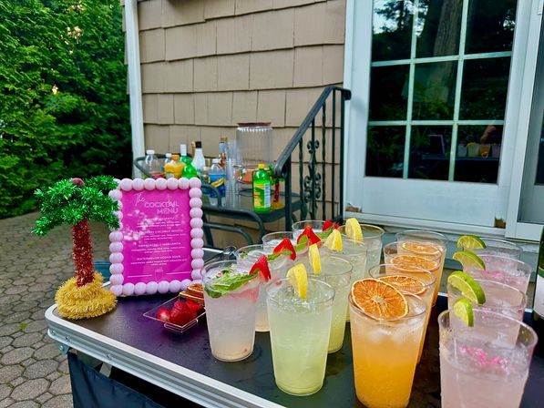 Outdoor backyard patio drink station with rows of colorful summer cocktails in plastic cups garnished with lemon wedges, lime slices, dried orange rounds, strawberries and mint, next to a pink-framed cocktail menu and a small decorative palm with mixers and house siding in the background.