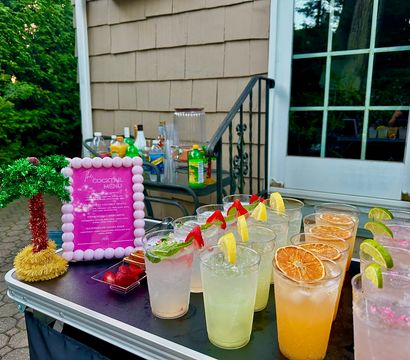 Outdoor backyard patio cocktail table with rows of colorful garnished drinks in plastic cups—lemon, lime, dried orange slices and strawberries—next to a pink-framed cocktail menu and a small tinsel palm tree, ready for a summer party.