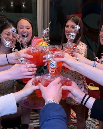 Friends toasting bright red cocktails with spiral disco-ball stirrers at an indoor celebration, smiling and clinking glasses.