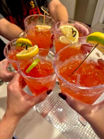 Four bright orange cocktails on ice with lemon and lime wedge garnishes, rimmed glasses raised in a cheerful toast at a party