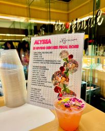 Bright rose-themed cocktail menu and a pink, rose-petal–garnished drink in a plastic cup on a mirrored bar counter beside a stack of disposable cups, event atmosphere