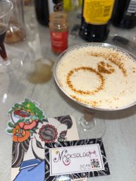 Cinnamon-dusted frothy cocktail in a martini glass with circular garnish on a bar countertop, surrounded by a spice jar, bottles, colorful floral skull stickers and a business card.