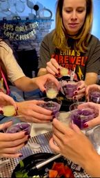 Close-up of friends clinking small plastic cups filled with purple and pale-yellow shots at an indoor farewell-style party, many hands with dark red nails and a festive backdrop.
