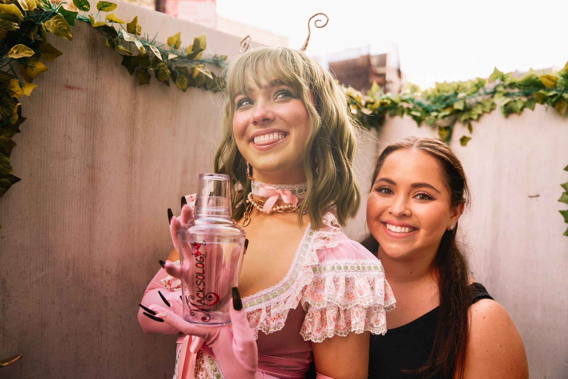 Two smiling women at an outdoor urban patio with ivy‑trimmed walls — one in a playful pink lace costume and gloves holding a clear cocktail shaker, the other in a black sleeveless top smiling behind her.