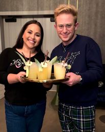 Two smiling people indoors holding a gold tray with four yellow pineapple cocktails garnished with pineapple slices and chocolate sticks — casual cocktail party scene.