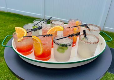 Cheerful tray of colorful summer cocktails with orange slices, lime and spiced rims on a white enamel tray set on a backyard lawn table