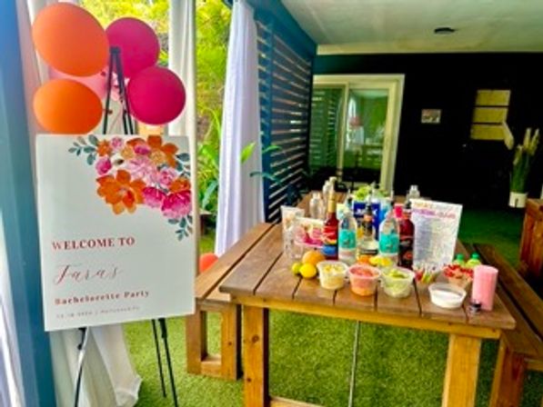 Covered patio bachelorette party setup with a floral welcome sign, pink and orange balloons, and a wooden table displaying bottles, mixers, lemons and small bowls of garnishes on artificial grass.