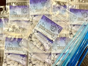 Flat lay of bridal party favor bags: clear resealable pouches with lavender watercolor custom labels and metallic confetti, arranged with bright blue plastic straws for wedding shower or bachelorette prep.