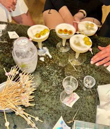 Overhead view of four frothy martini-style cocktails on a granite bar, each garnished with three skewered pearl onions; cocktail shaker, pile of drink picks, shot glasses and hands nearby for a casual bar night vibe.