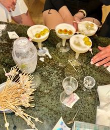 Four frothy coupe cocktails garnished with skewered green olives on a dark granite bar countertop, surrounded by a cocktail shaker and a pile of decorative skewers.
