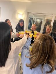 Indoor house party toast: group of women smiling and clinking bright yellow cocktails in martini glasses around a kitchen island.