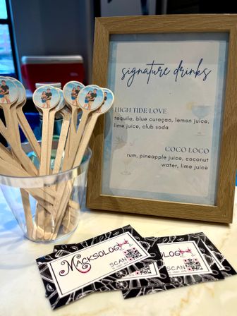 Framed “signature drinks” cocktail menu beside a clear cup of wooden stir sticks with round photo toppers and stacked promo cards on a marble countertop — casual indoor event bar display.