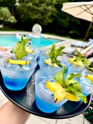 Tray of vibrant blue cocktails with ice, mint sprigs and starfruit garnishes, served poolside at a sunny backyard swimming pool with lounge chairs and umbrella in the background.