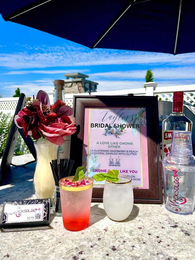 Sunny outdoor patio bridal shower drink station with framed bridal shower sign, pink and white garnish cocktails topped with rose petals and basil, burgundy floral centerpiece, liquor bottle and cocktail shaker on a granite table under a navy umbrella.