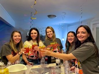 Five friends in a home kitchen toasting garnished cocktails, wearing matching farewell-tour T-shirts beneath hanging spiral streamers.