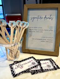 Playful cocktail station close-up: wooden drink stirrers with round photo toppers in a clear cup beside a framed “signature drinks” menu listing High Tide Love and Coco Loco, with stacked promotional cards on a marble countertop.