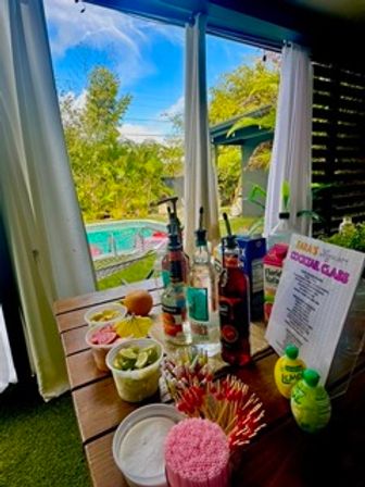 Poolside DIY cocktail station on a wooden patio table framed by white curtains, with liquor bottles, citrus garnishes, skewers and a mix-your-own menu overlooking a backyard pool and lush greenery.