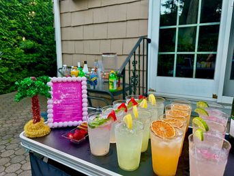 Backyard patio cocktail station with colorful mixed drinks in clear cups—garnished with lemon, lime, strawberries and dried orange—beside a pink-framed menu and a small tinsel palm.