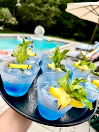 Vibrant blue cocktails on a black tray with crushed ice, starfruit slices and fresh mint, set poolside with a blurred backyard pool and umbrella in the background — tropical summer drinks.