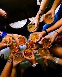 Overhead shot of a group toast — hands holding colorful cocktails garnished with lime wedges, cherries and rose petals, many wrists wearing bracelets at an indoor summer gathering.