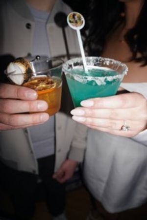Close-up of two hands holding cocktails at a bar: a blue sugar-rimmed cocktail with stirrer and an amber cocktail with orange garnish, lively nightlife scene