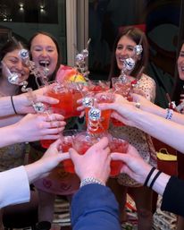 Group of friends cheering with red cocktails in clear glasses, spiral stirrers topped with mini disco-ball charms, laughing at an indoor party celebration.