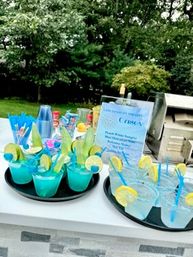Two black trays of bright blue cocktails and clear mocktails in plastic cups with lime slices and blue straws on a white table at a backyard grill party with trees in the background.