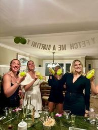 Four women in a home kitchen raising neon green cocktail shakers and smiling under a party banner during a bachelorette-style celebration.