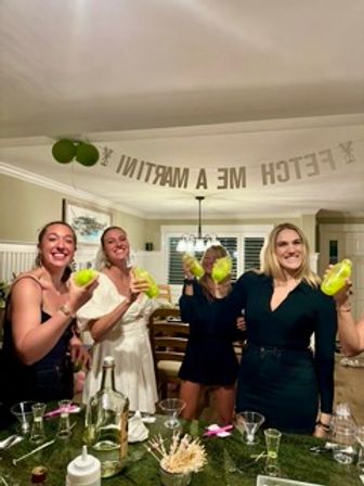 Four women in a home kitchen raising neon green cocktail shakers and smiling under a party banner during a bachelorette-style celebration.