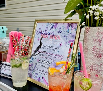 Backyard bridal shower drink station with framed floral cocktail menu, three colorful iced cocktails garnished with citrus, lavender and jalapeño, floral vase and bright paper straws