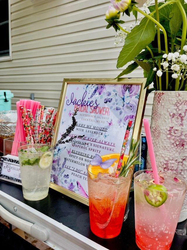 Backyard bridal shower drink station with framed floral cocktail menu, three colorful iced cocktails garnished with citrus, lavender and jalapeño, floral vase and bright paper straws