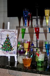 Bright holiday cocktail display: multicolored flute tower and mixed cocktails on a bar countertop next to a festive Christmas-tree menu