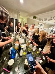 Friends gathered around a home kitchen island mixing cocktails and celebrating a bachelorette party — bride in a veil chats while others take photos.
