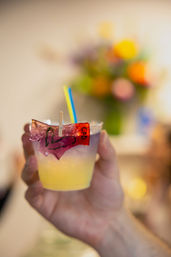 Hand holding a small clear cup with a yellow iced cocktail, blue-and-yellow straw and a playful purple candy garnish clipped to the rim, blurred colorful bouquet in the background.