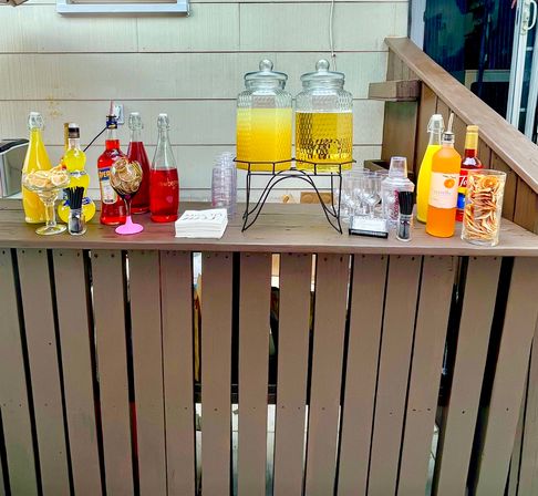 Cheerful backyard patio bar on a wooden deck railing with two large glass beverage dispensers of orange and yellow drinks, colorful bottles and mixers, glassware, napkins and citrus garnishes arranged for a casual outdoor party.