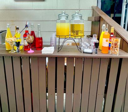 Cheerful backyard patio bar on a wooden deck railing with two large glass beverage dispensers of orange and yellow drinks, colorful bottles and mixers, glassware, napkins and citrus garnishes arranged for a casual outdoor party.
