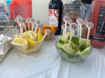 Glass bowls of lemon, lime, and orange wedges with wooden cocktail picks topped with small round photo stickers, arranged on a plastic-covered table with bottles of spirits and bar tools in the background.