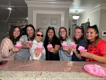 Seven friends smiling behind a granite kitchen counter, each holding a pink-frosted cupcake at a casual home gathering.