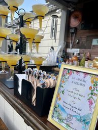 Tower of sunny yellow cocktails in coupe glasses on a metal stand, beside wooden stirrers and a framed floral drink menu at an outdoor bar.