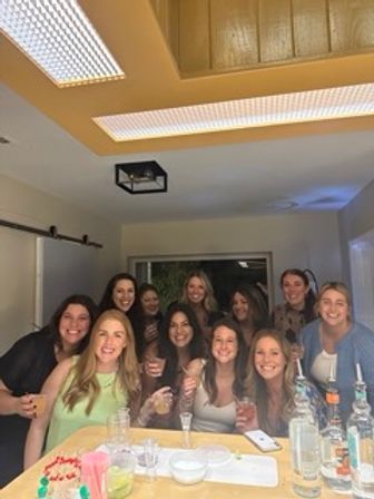 Smiling group of women gathered around a kitchen island with cocktails, bottles and snacks, enjoying a lively indoor house party.