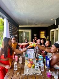 Cheerful friends toasting with colorful cocktails around a rustic wooden table on a covered patio—brunch-style summer gathering with fruit, mixers and glassware.