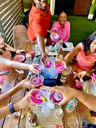 Friends toasting bright pink cocktails around a wooden outdoor-patio table at a sunny brunch, garnished drinks and bottles on the table.