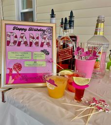 Cheerful pink-framed birthday sign and drink station on a porch table with bottles of spirits, two colorful cocktails garnished with lime, pink stirrers and party picks.