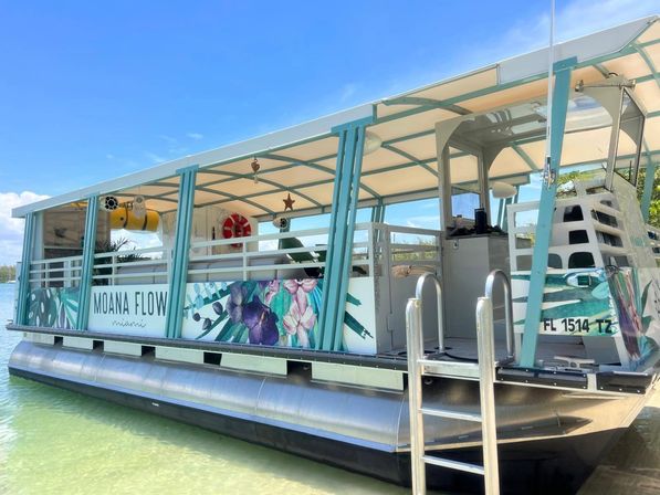 Cheerful turquoise-and-white covered pontoon boat with floral panels and life rings, moored in clear shallow coastal Florida waters under a bright blue sky.