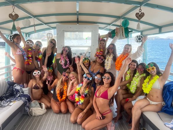 Smiling women in bikinis, sunglasses and colorful leis posing and cheering on a covered boat party with a coastal city skyline in the background.