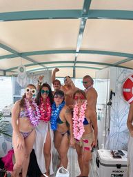 Summer boat party on a covered pontoon — group of adults in swimsuits, colorful leis and fun sunglasses posing near a lake with drinks and a cooler