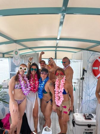Summer boat party on a covered pontoon — group of adults in swimsuits, colorful leis and fun sunglasses posing near a lake with drinks and a cooler